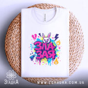 Футболка зла зая, білого кольору, складена на плетеній кошику, фон світло-сірий, принт яскравий, з зображенням злого зайця та кольоровими сплесками фарби, видно етикетку.