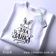 Футболка зла зайка, біла, на столі, з зеленим ярликом, фон світлий, принт з зображенням злого зайця та написом 'ЗЛА ЗАЙКА' у чорному кольорі, вид зверху.