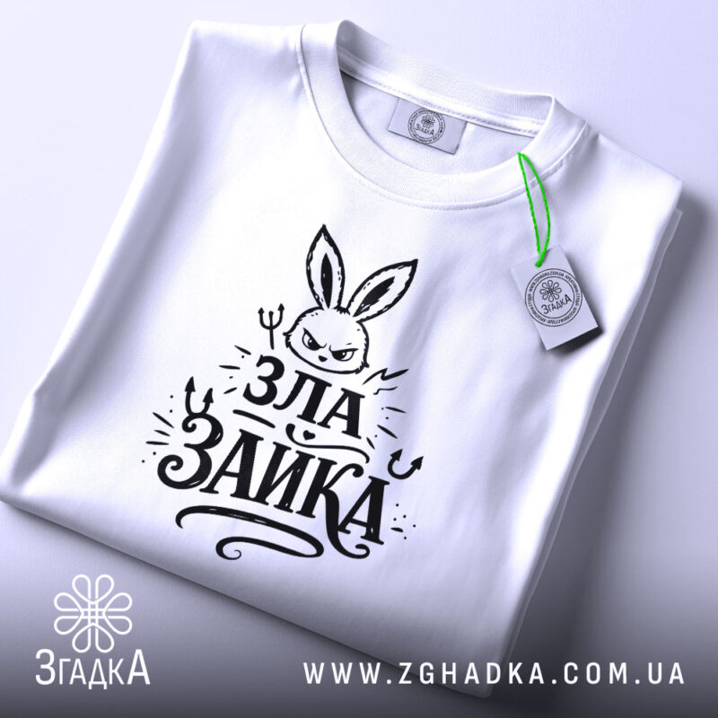 Футболка зла зайка, біла, на столі, з зеленим ярликом, фон світлий, принт з зображенням злого зайця та написом 'ЗЛА ЗАЙКА' у чорному кольорі, вид зверху.