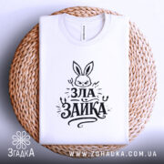 Футболка зла зайка, біла, на плетеній кошику, фон світлий, принт з зображенням злого зайця та написом 'ЗЛА ЗАЙКА' у чорному кольорі, вид зверху.