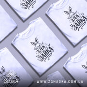 Футболка зла зайка, біла, на моделі, з легким макіяжем та волоссям, що спадає на плече, фон світло-фіолетовий, принт з зображенням злого зайця з великими вухами та написом 'ЗЛА ЗАЙКА' у чорному кольорі.