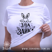 Футболка зла зайка, біла, на столі, з зеленим ярликом, фон світлий, принт з зображенням злого зайця та написом 'ЗЛА ЗАЙКА' у чорному кольорі, вид зверху.