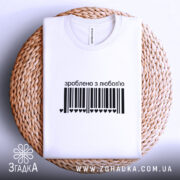 Футболка зроблено з любов’ю, біла, на круглому кошику, з чорним принтом у вигляді штрих-коду та написом 'зроблено з любов’ю', фон світло-сірий.