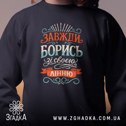 Світшот борись зі своєю лінню, чорного кольору, з круглою горловиною, близький план на принт з написом 'Завжди борись зі своєю лінню', манжети та трикотажна резинка на поясі, фактура м'яка, фон розмитий.