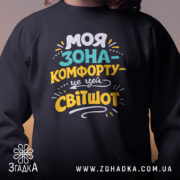 Світшот моя зона комфорту, чорного кольору, з круглою горловиною, з близького ракурсу на моделі, фон нейтральний, принт з яскравими кольорами, манжети та трикотажна резинка на поясі, фактура м'яка та комфортна.