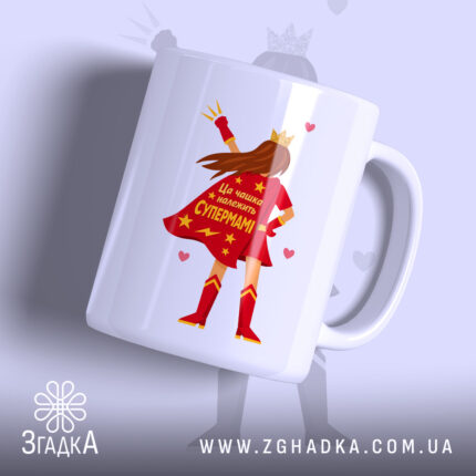 Чашка для Супермами, біла, зображена з легким нахилом, на фоні світло-фіолетового кольору, з принтом супергероїні в червоному плащі, на якому написано 'Ця чашка належить Супермамі', ручка класична, край рівний, глянцевий.