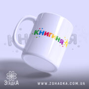 Чашка подарунок для книголюба, біла, з легким блиском, з написом 'КНИГИНЯ' кольоровими літерами, ручка класична, край рівний, чашка нахилена.