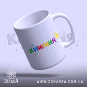 Чашка подарунок для книголюба, біла, з легким блиском, з написом 'КНИГИНЯ' кольоровими літерами, ручка класична, край рівний, чашка нахилена.