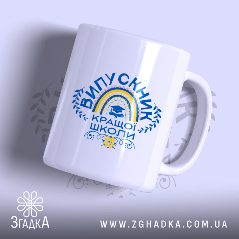 Чашка випускник кращої школи, зображена з легкого ракурсу, на світлому фоні, має яскравий принт з написом 'ВИПУСКНИК КРАЩОЇ ШКОЛИ' та веселкою, ручка круглої форми, край чашки рівний, глянцева поверхня.