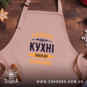 Фартух Бабуся на кухні, бежевого кольору, зображений на дерев'яному фоні, ракурс зверху, принт з текстом 'У Бабусі на кухні завжди смачно' в темно-синьому та жовтому кольорах, поряд з декором у вигляді сніжинки та печива.