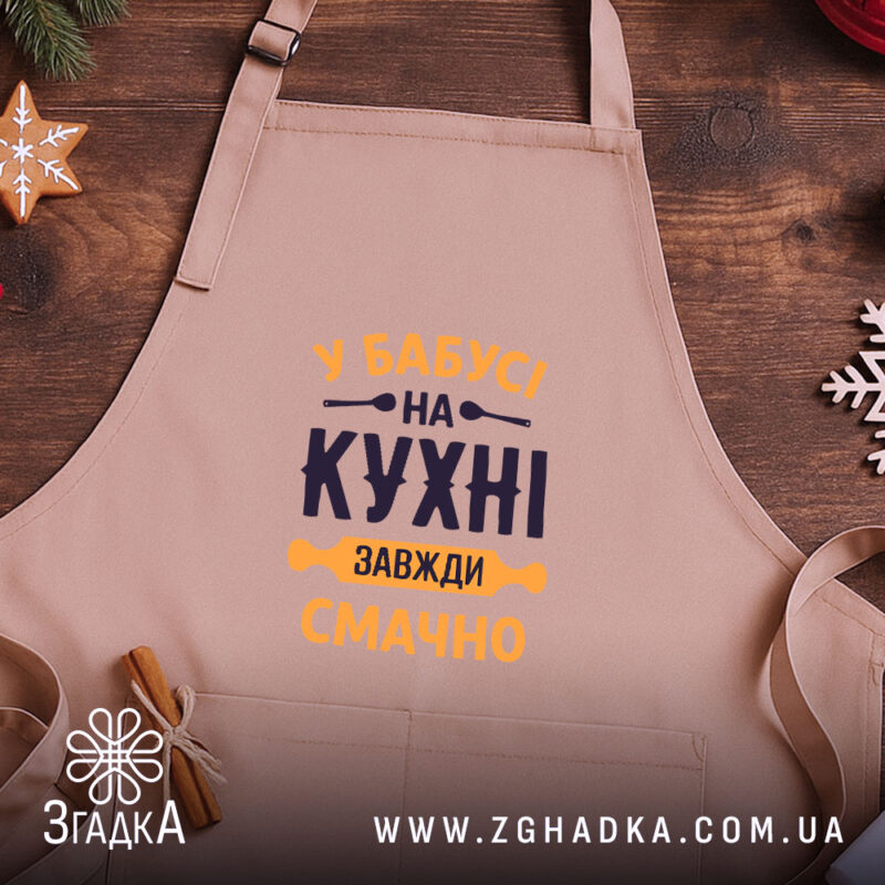Фартух Бабуся на кухні, бежевого кольору, зображений на дерев'яному фоні, ракурс зверху, принт з текстом 'У Бабусі на кухні завжди смачно' в темно-синьому та жовтому кольорах, поряд з декором у вигляді сніжинки та печива.