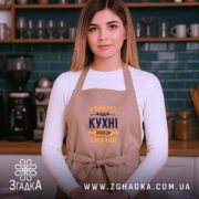 Фартух Бабуся на кухні, бежевого кольору, зображений на жінці, яка стоїть у кухні, ракурс спереду, фон з кухонними приладами, принт з текстом 'У Бабусі на кухні завжди смачно' в темно-синьому та жовтому кольорах.
