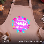 Фартух головна мама країни, бежевого кольору, зображений на дерев'яному фоні, деталі принта з квітами та написом 'Головна мама країни'.