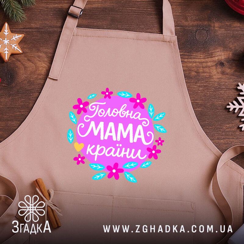 Фартух головна мама країни, бежевого кольору, зображений на дерев'яному фоні, деталі принта з квітами та написом 'Головна мама країни'.