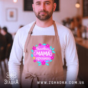 Фартух головна мама країни, бежевого кольору, на чоловікові, фон кафе з розмитими елементами, принт з квітами та написом 'Головна мама країни'.