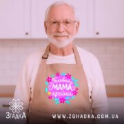 Фартух головна мама країни, бежевого кольору, зображений на дерев'яному фоні, деталі принта з квітами та написом 'Головна мама країни'.