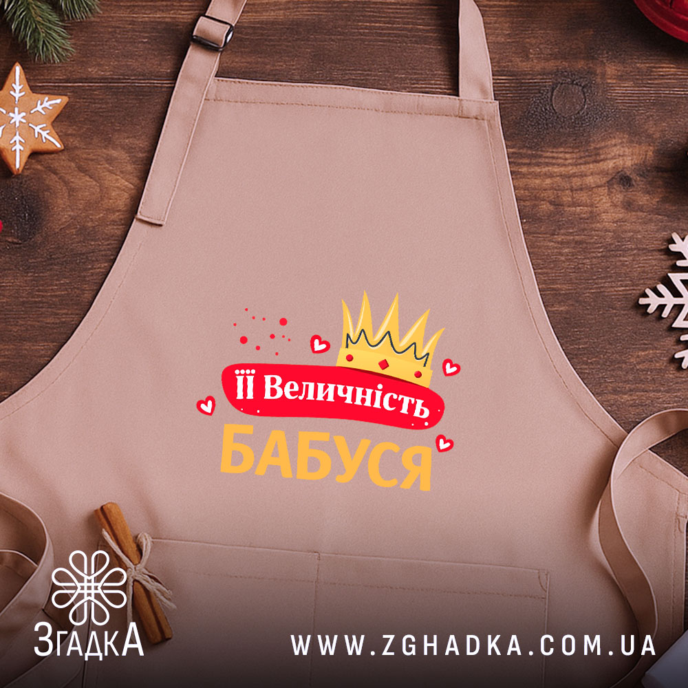 Фартух її величність бабуся, бежевого кольору, зображений зверху на дерев'яному фоні, принт з короною та сердечками, напис червоним і жовтим кольором, поруч з корицею та новорічними прикрасами.