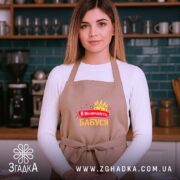 Фартух її величність бабуся, бежевого кольору, зображений на жінці з довгим волоссям, фон кухонний з темно-синьою стіною, принт з короною та сердечками, напис червоним і жовтим кольором.