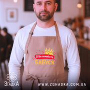 Фартух її величність бабуся, бежевого кольору, зображений на чоловікові з бородою, фон кафе з розмитими людьми, принт містить корону та сердечка, напис червоним і жовтим кольором.