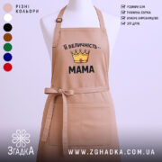 Фартух Її Величність Мама бежевого кольору, зображений з фронтального ракурсу, на фоні світлого однотонного фону. На фартусі є принт у вигляді корони жовтого кольору з червоним каменем та написом "Її Величність – МАМА" чорним шрифтом.