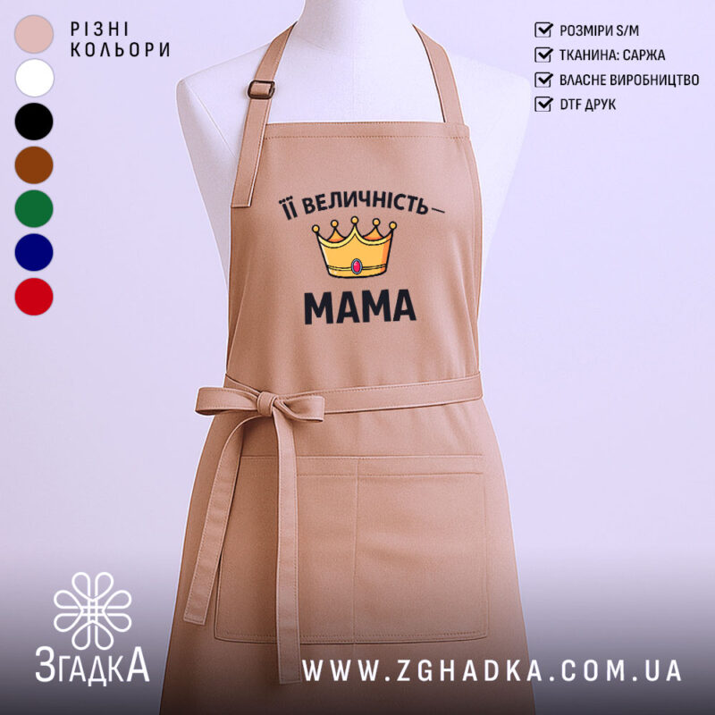 Фартух Її Величність Мама бежевого кольору, зображений з фронтального ракурсу, на фоні світлого однотонного фону. На фартусі є принт у вигляді корони жовтого кольору з червоним каменем та написом "Її Величність – МАМА" чорним шрифтом.