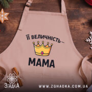 Фартух Її Величність Мама бежевого кольору, зображений з верхнього ракурсу на дерев'яному фоні. На фартусі розташований принт у вигляді корони жовтого кольору з червоним каменем та написом "Її Величність – МАМА" чорним шрифтом, поруч видно декор у вигляді новорічних прикрас.