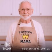 Фартух Її Величність Мама бежевого кольору, зображений з верхнього ракурсу на дерев'яному фоні. На фартусі розташований принт у вигляді корони жовтого кольору з червоним каменем та написом "Її Величність – МАМА" чорним шрифтом, поруч видно декор у вигляді новорічних прикрас.