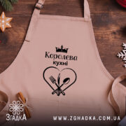 Фартух королева кухні, бежевого кольору, зображений на дерев'яному фоні, деталі принта чорного кольору з написом 'Королева кухні' та зображенням серця з виделкою і ложкою, поруч розташовані святкові елементи.