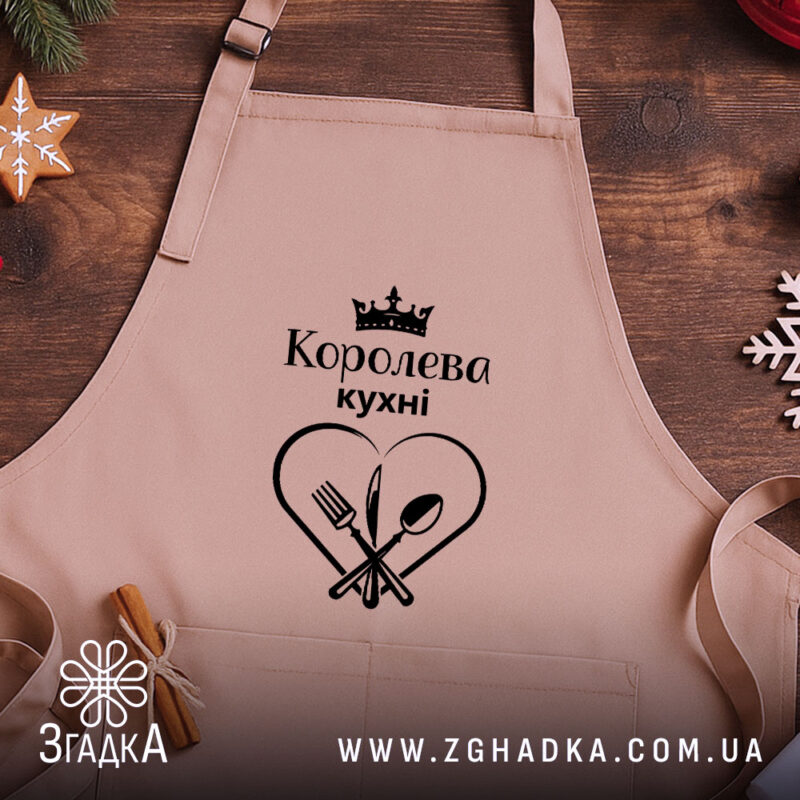 Фартух королева кухні, бежевого кольору, зображений на дерев'яному фоні, деталі принта чорного кольору з написом 'Королева кухні' та зображенням серця з виделкою і ложкою, поруч розташовані святкові елементи.