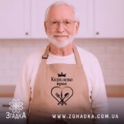 Фартух королева кухні, бежевого кольору, зображений на дерев'яному фоні, деталі принта чорного кольору з написом 'Королева кухні' та зображенням серця з виделкою і ложкою, поруч розташовані святкові елементи.