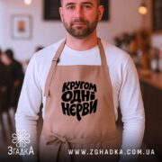 Фартух кругом одні нєрви, бежевий на чоловікові, фон кафе, стильний крій, бренд.