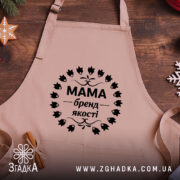 Фартух Мама бежевого кольору, зображений зверху на дерев'яному столі, фон натуральний, деталі принта чорного кольору з написом 'Мама бренд якості' в оточенні листя, поруч розташовані кухонні аксесуари.