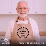 Фартух Мама бежевого кольору, зображений зверху на дерев'яному столі, фон натуральний, деталі принта чорного кольору з написом 'Мама бренд якості' в оточенні листя, поруч розташовані кухонні аксесуари.