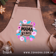 Фартух мама найкраща у світі, бежевого кольору, зображений на дерев'яному фоні, ракурс зверху, принт з квітами та написом 'Найкраща МАМА у світі'.