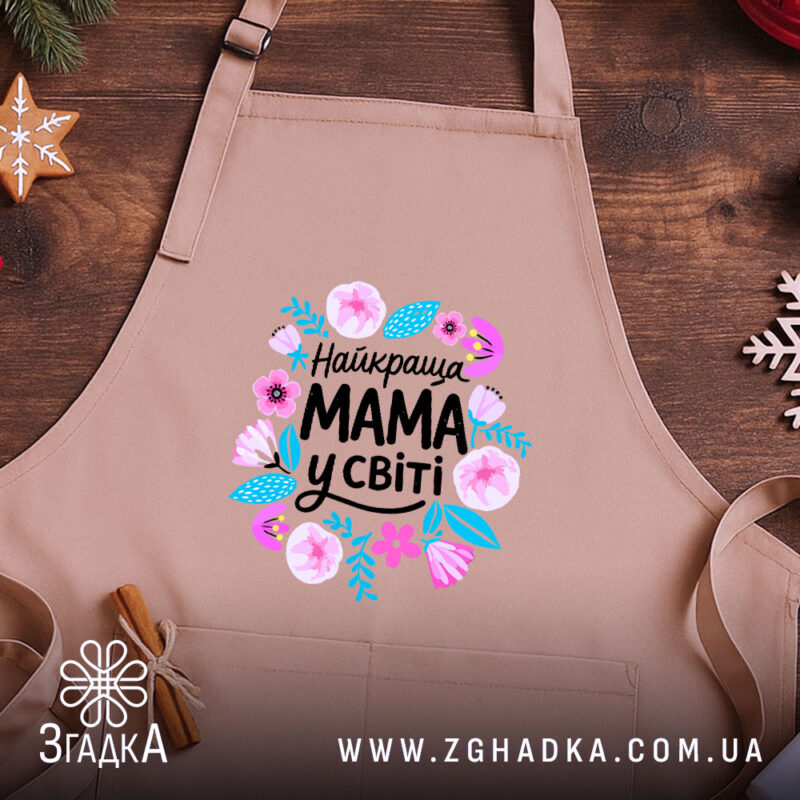 Фартух мама найкраща у світі, бежевого кольору, зображений на дерев'яному фоні, ракурс зверху, принт з квітами та написом 'Найкраща МАМА у світі'.