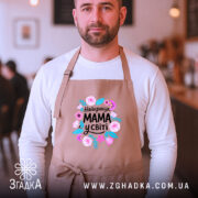 Фартух мама найкраща у світі, бежевого кольору, на чоловікові, фон кафе, принт з квітами та написом 'Найкраща МАМА у світі'.