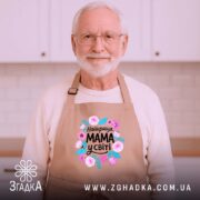 Фартух мама найкраща у світі, бежевого кольору, зображений на дерев'яному фоні, ракурс зверху, принт з квітами та написом 'Найкраща МАМА у світі'.