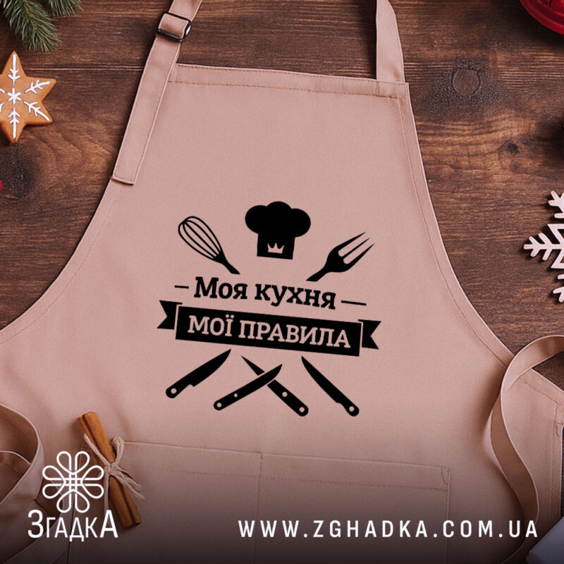 Фартух моя кухня мої правила. Бежевий фартух, знятий з близького ракурсу на дерев'яному фоні. На фоні видно декор у вигляді новорічних зірочок. Принт чорного кольору з написом 'Моя кухня мої правила' та зображеннями кухонних інструментів.
