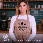 Фартух моя кухня мої правила. Бежевий фартух, зображений на жінці в кухні, знімок з фронтального ракурсу. Фон складається з кухонних полиць з посудом. Принт чорного кольору з написом 'Моя кухня мої правила' та зображеннями кухонних інструментів.