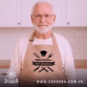 Фартух моя кухня мої правила. Бежевий фартух, знятий з близького ракурсу на дерев'яному фоні. На фоні видно декор у вигляді новорічних зірочок. Принт чорного кольору з написом 'Моя кухня мої правила' та зображеннями кухонних інструментів.