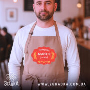 Фартух найкраща бабуся, бежевого кольору, на чоловікові з бородою, фон кафе з розмитими людьми, принт з написом 'Найкраща Бабуся у світі' в червоній рамці.