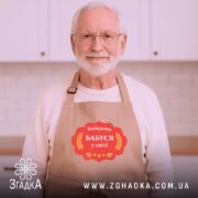 Фартух найкраща бабуся, бежевого кольору, зображений на дерев'яному фоні, деталі принта аналогічні, на фоні присутні святкові елементи.