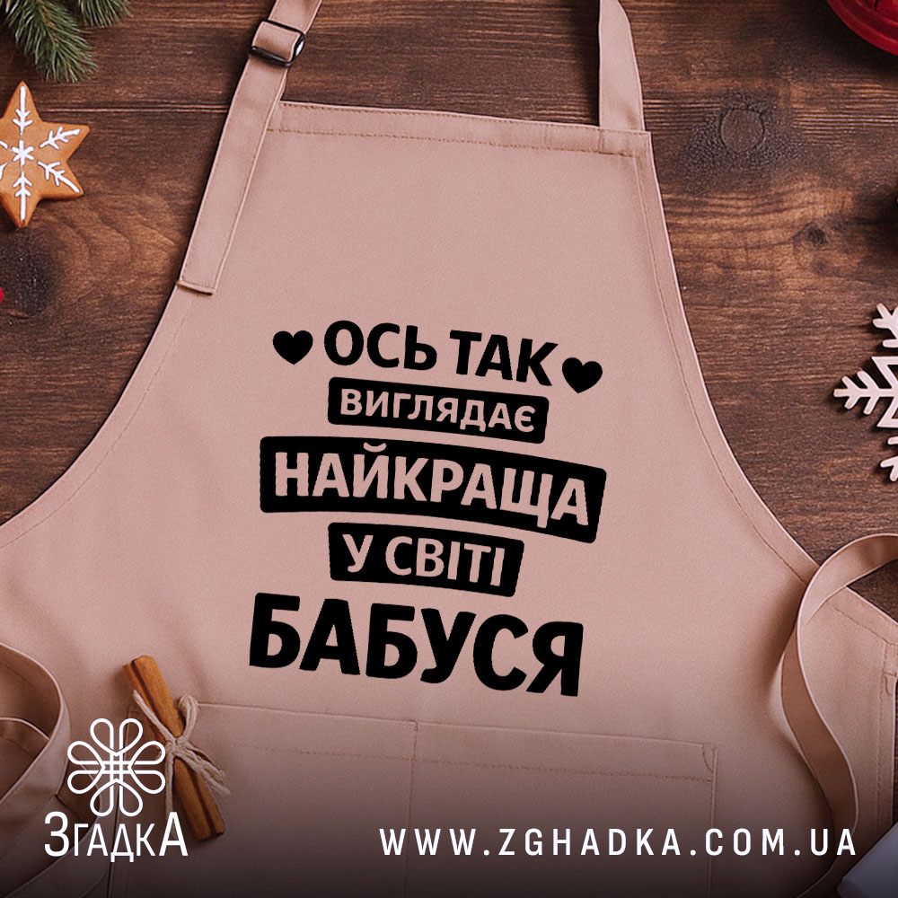 Фартух найкраща бабуся, бежевого кольору, зображений на дерев'яному фоні, принт чорного кольору з текстом 'Ось так виглядає найкраща у світі бабуся', на фоні присутні святкові елементи.