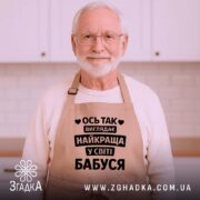 Фартух найкраща бабуся, бежевого кольору, зображений на дерев'яному фоні, принт чорного кольору з текстом 'Ось так виглядає найкраща у світі бабуся', на фоні присутні святкові елементи.