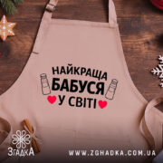 Фартух найкраща бабуся, бежевого кольору, зображений з верхнього ракурсу на дерев'яному фоні, деталі принта з текстом "НАЙКРАЩА БАБУСЯ У СВІТІ" на фоні святкових елементів.
