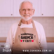 Фартух найкраща бабуся, бежевого кольору, зображений з верхнього ракурсу на дерев'яному фоні, деталі принта з текстом "НАЙКРАЩА БАБУСЯ У СВІТІ" на фоні святкових елементів.