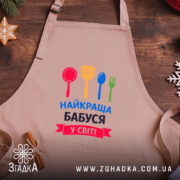 Фартух найкраща бабуся у світі, бежевого кольору, розташований на дерев'яному фоні, принт з червоним, жовтим, зеленим та синім кухонним приладдям, навколо декор у вигляді новорічних зірочок.