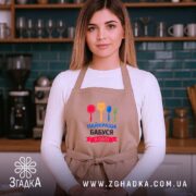 Фартух найкраща бабуся у світі, бежевого кольору, на моделі з довгим волоссям, фон темно-синій з кухонними приладами, принт містить червоний, жовтий, зелений та синій кольори кухонного приладдя.