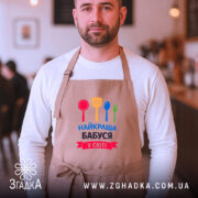 Фартух найкраща бабуся у світі, бежевого кольору, на чоловікові з бородою, фон розмитий, з елементами кафе, принт з червоним, жовтим, зеленим та синім кухонним приладдям.