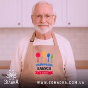 Фартух найкраща бабуся у світі, бежевого кольору, розташований на дерев'яному фоні, принт з червоним, жовтим, зеленим та синім кухонним приладдям, навколо декор у вигляді новорічних зірочок.